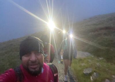 a man smiling at the camera with head torches glaring in the background climing up mount snowden