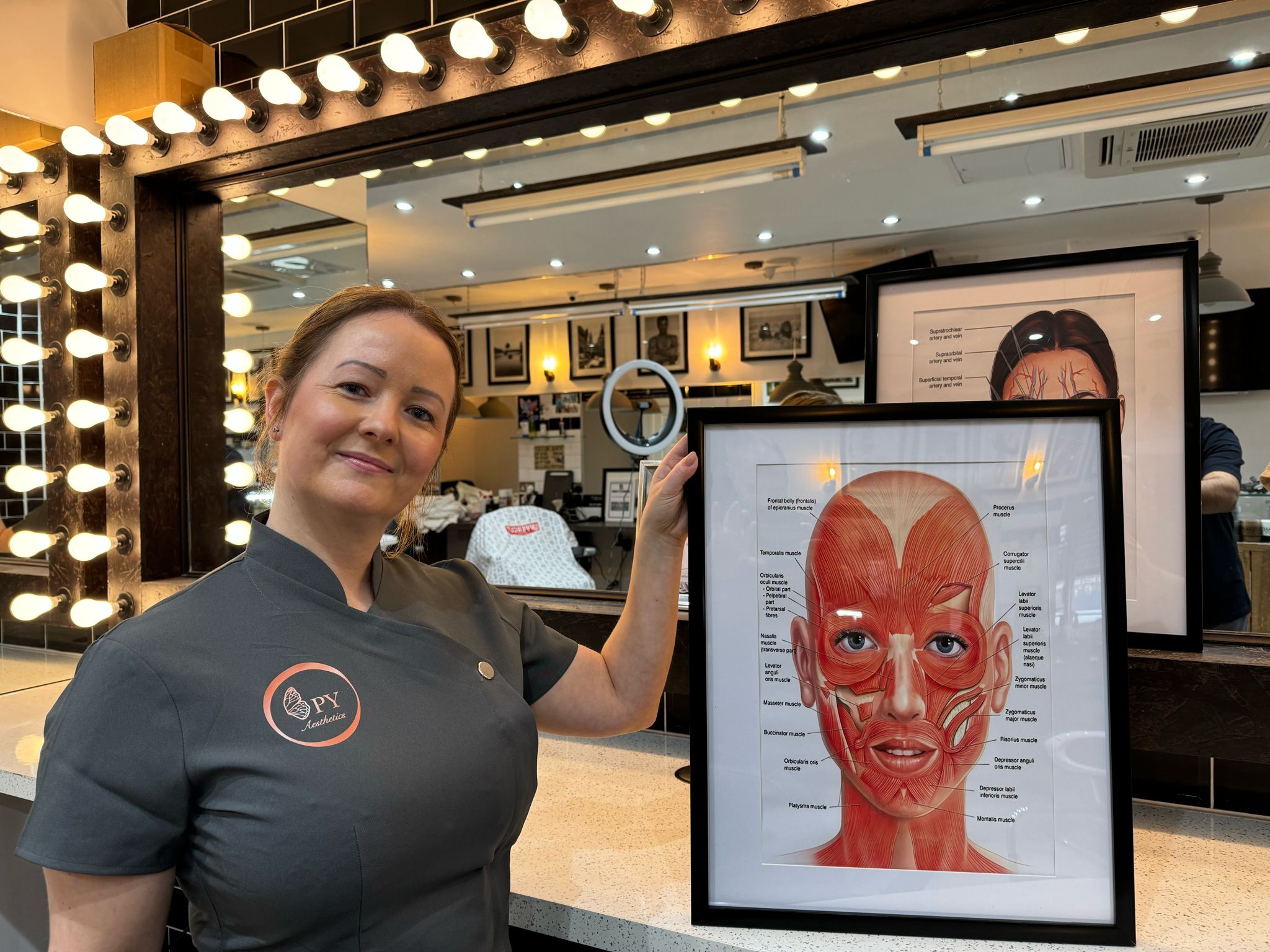 Lady holding a framed medical image of the muscles of the face smiling at the camera in seven hair and body barbers in peterborough